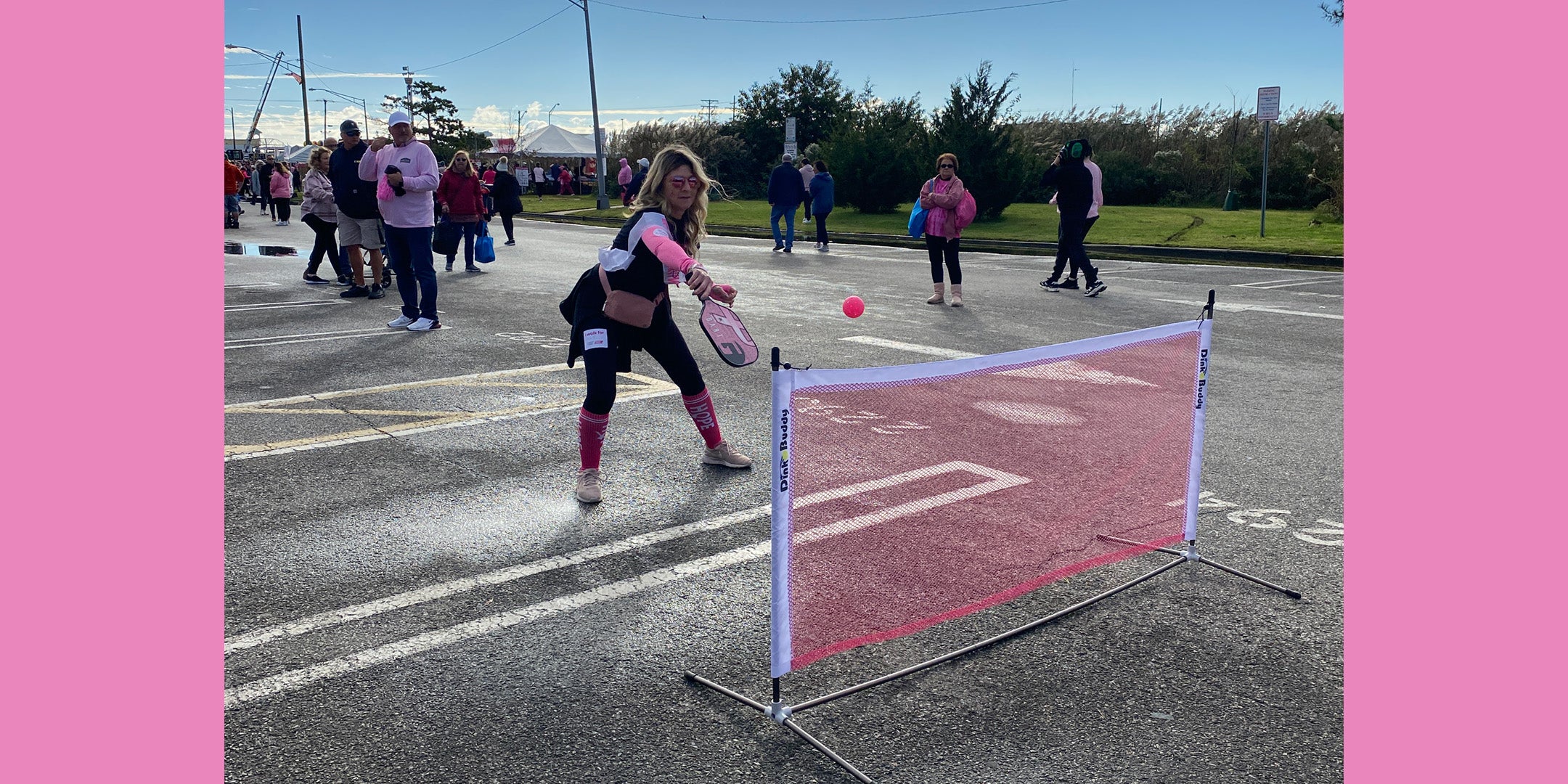 A woman in New Jersey plays pickleball with a pink paddle and ball on a portable net in a parking lot during a breast cancer awareness event, surrounded by participants wearing pink.